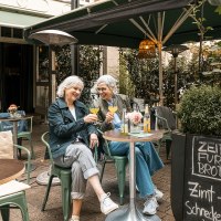 Zwei Frauen sitzen in einem gemütlichen Café im Freien, stoßen mit Getränken an und lachen. Ein Schild bietet Zimtschnecken an., © Stuttgart Marketing GmbH - Sarah Schmid Zwei Frauen sitzen in einem gemütlichen Café im Freien, stoßen mit Getränken an und lachen. Ein Schild bietet Zimtschnecken an., © Stuttgart Marketing GmbH - Sarah Schmid