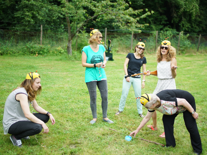 Frauen mit Bienenhüten spielen ein Outdoor-Spiel auf einer Wiese. Eine Frau kniet, während andere stehen und lachen., © Cool-Tours StattReisen Frauen mit Bienenhüten spielen ein Outdoor-Spiel auf einer Wiese. Eine Frau kniet, während andere stehen und lachen., © Cool-Tours StattReisen