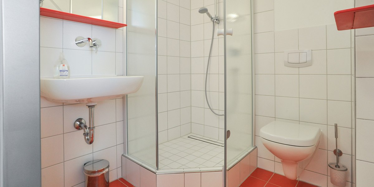 A modern bathroom with white-tiled walls, a shower, a sink, and a toilet. Red accents and a trash can are visible., &copy; G&auml;stehaus ABZ Stuttgart