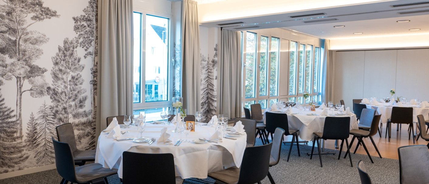Elegant breakfast room with round, white-clothed tables, modern chairs and large windows. One wall is decorated with tree illustrations., © Erikson Hotel