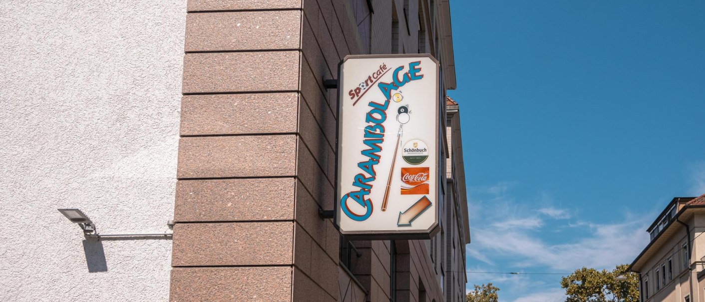 Ein Schild mit der Aufschrift 'Carambolage' hängt an einer Gebäudewand. Der Himmel ist klar und blau., © SMG, Sarah Schmid Ein Schild mit der Aufschrift 'Carambolage' hängt an einer Gebäudewand. Der Himmel ist klar und blau., © SMG, Sarah Schmid