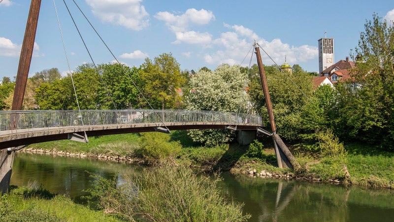 A modern pedestrian bridge spans a river, surrounded by green vegetation. A church tower can be seen in the background., © Stuttgart-Marketing GmbH A modern pedestrian bridge spans a river, surrounded by green vegetation. A church tower can be seen in the background., © Stuttgart-Marketing GmbH