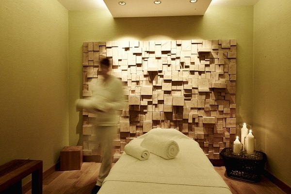 A massage room with a wooden decorative wall, massage table and candles at the Waldhotel Stuttgart. A person moves around the room., &copy; Waldhotel Stuttgart GmbH