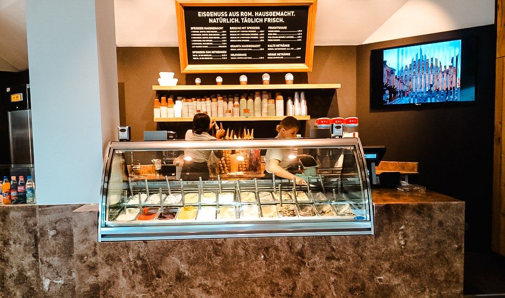 Eisdiele mit Theke, gefüllt mit verschiedenen Eissorten. Zwei Personen arbeiten dahinter. An der Wand ein Menü und ein Bildschirm., © Old Bridge Gelateria Stuttgart