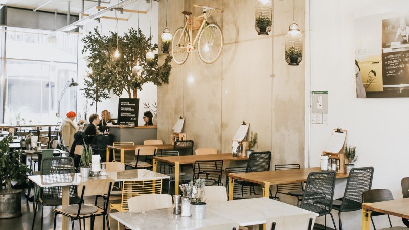 Modernes Café mit hängendem Fahrrad an der Wand, Pflanzen und gemütlichen Sitzbereichen. Menschen sitzen und unterhalten sich an Tischen., © Claus Stuttgart Modernes Café mit hängendem Fahrrad an der Wand, Pflanzen und gemütlichen Sitzbereichen. Menschen sitzen und unterhalten sich an Tischen., © Claus Stuttgart