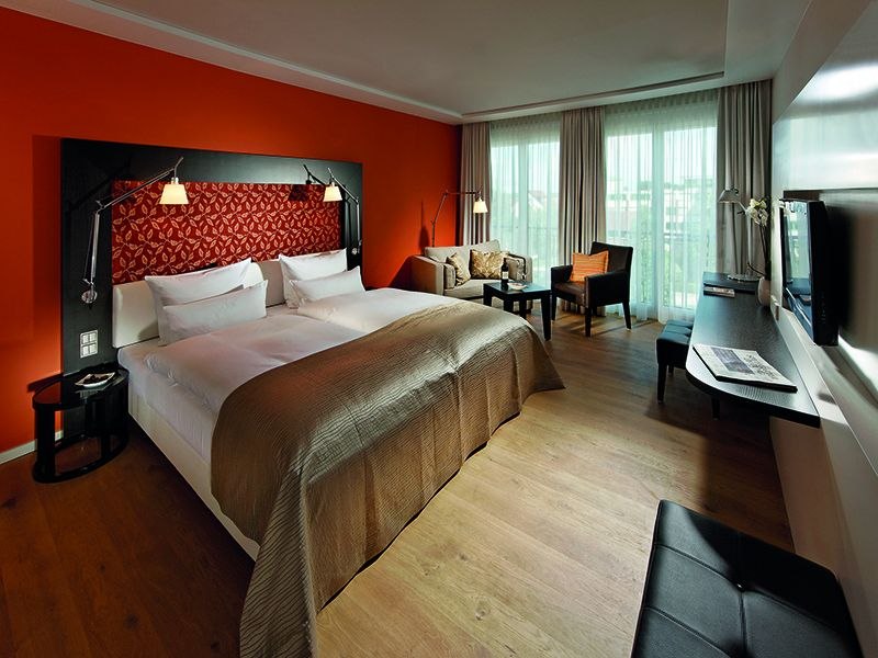 Modernes Hotelzimmer mit gro&szlig;em Bett, oranger Wand, Sofa, Schreibtisch und Fernseher. Helle Vorh&auml;nge und Holzboden schaffen eine gem&uuml;tliche Atmosph&auml;re., &copy; Parkhotel Stuttgart Messe-Airport