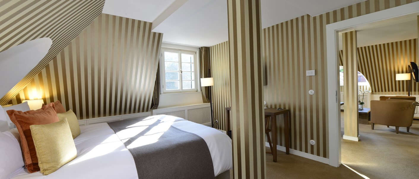 Elegante Hotelsuite mit gestreiften Wänden, gemütlichem Bett und angrenzendem Wohnzimmer. Helle Fenster und stilvolle Einrichtung schaffen eine einladende Atmosphäre., © Waldhotel Stuttgart GmbH