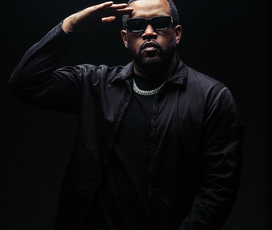 Eine Person in schwarzer Kleidung und Sonnenbrille salutiert vor einem dunklen Hintergrund. Die Beleuchtung hebt die Person hervor., &copy; SBEntertainment GmbH & Co. KG