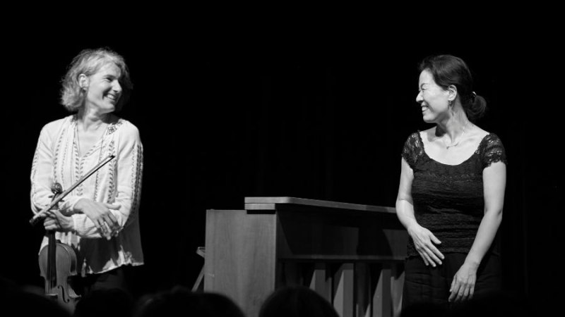 Zwei Frauen auf einer Bühne, eine mit Geige, die andere am Klavier, lächeln einander an. Schwarz-Weiß-Foto., © Fotocredit Theater La Lune Stuttgart