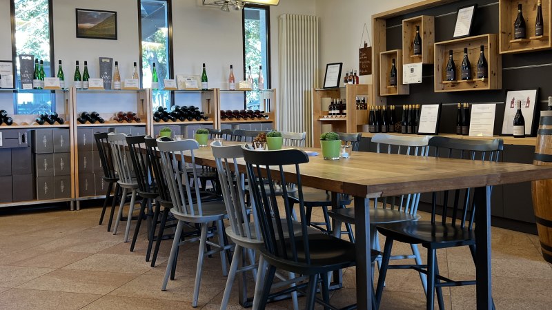 Vinothek mit Holztisch und Stühlen, umgeben von Weinregalen und Dekoration. Helle, einladende Atmosphäre mit Weinflaschen und Pflanzen., © Weinfactum Bad Cannstatt Vinothek mit Holztisch und Stühlen, umgeben von Weinregalen und Dekoration. Helle, einladende Atmosphäre mit Weinflaschen und Pflanzen., © Weinfactum Bad Cannstatt