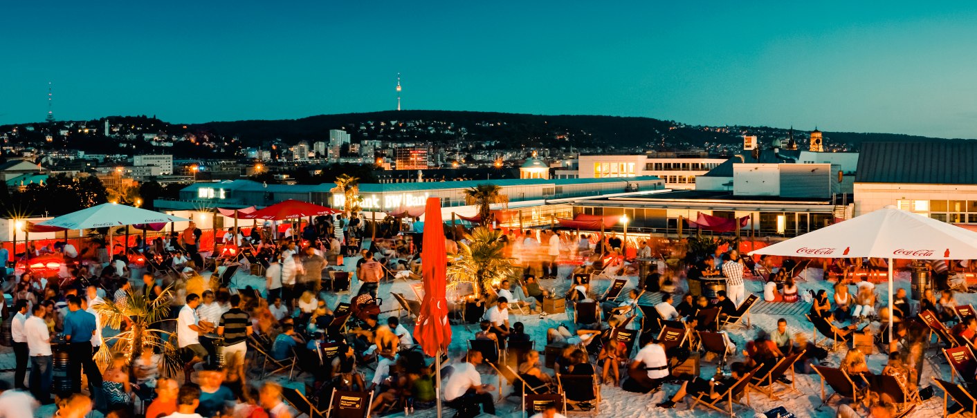 Große Menschenmenge auf einer Dachterrasse bei Abenddämmerung, umgeben von Sonnenschirmen und Liegestühlen, mit Blick auf eine beleuchtete Stadt., © Sky Beach Große Menschenmenge auf einer Dachterrasse bei Abenddämmerung, umgeben von Sonnenschirmen und Liegestühlen, mit Blick auf eine beleuchtete Stadt., © Sky Beach
