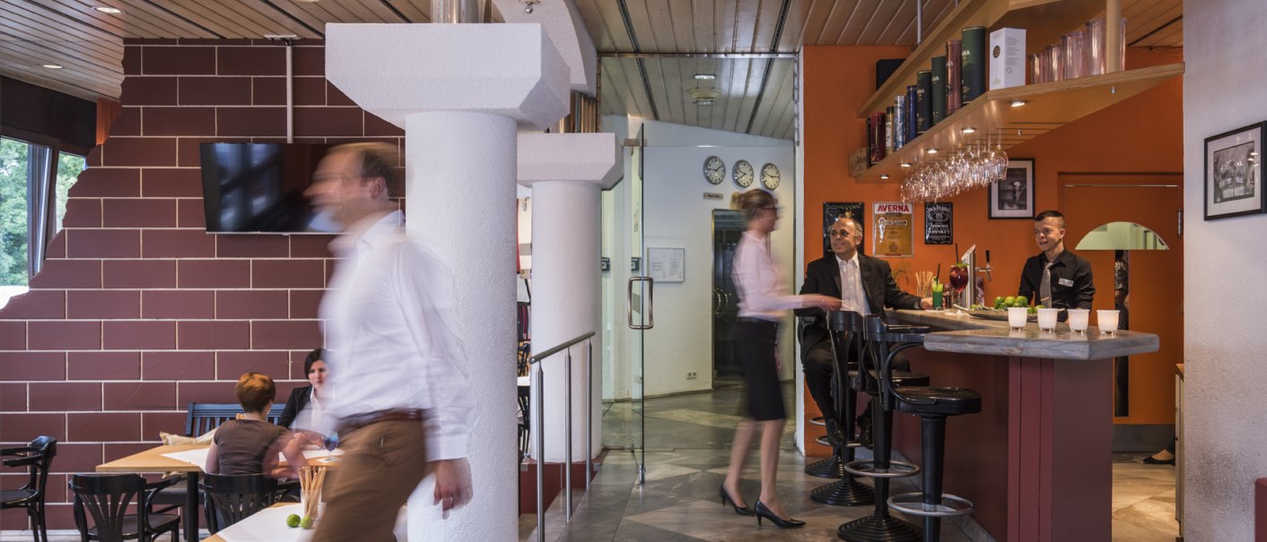 Ein Bistro mit Bar, an der zwei Männer sitzen. Eine Frau geht vorbei. Im Hintergrund sitzen Gäste an Tischen. Die Atmosphäre ist lebhaft., © Hotel Berlin aZIS Hotel BetriebsGmbH