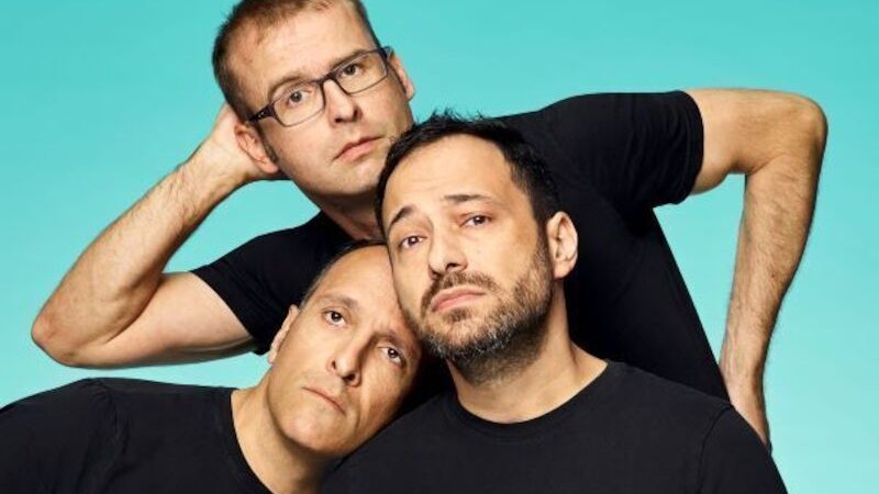 Three men in black T-shirts pose in front of a turquoise background. They look serious and are leaning close together., &copy; Rosenau Kultur e.V.