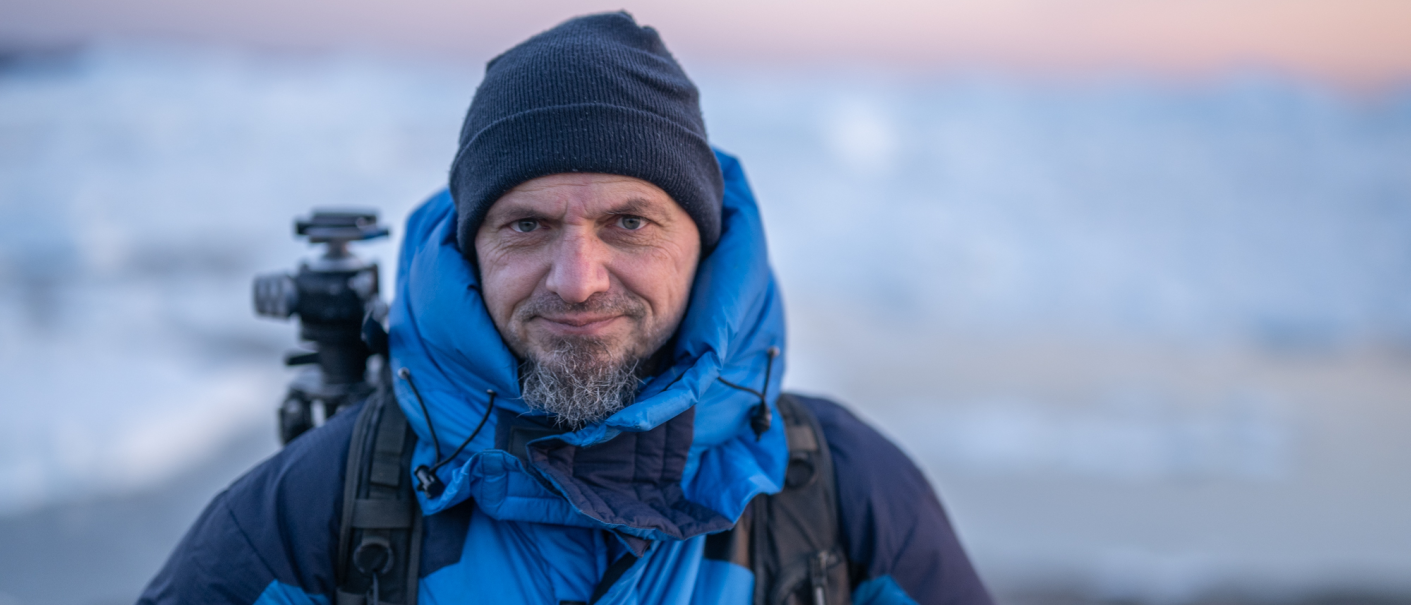 A man in a blue winter jacket and cap stands in front of a snow-covered landscape. He is carrying a rucksack with camera equipment., &copy; Andr&eacute; Delia