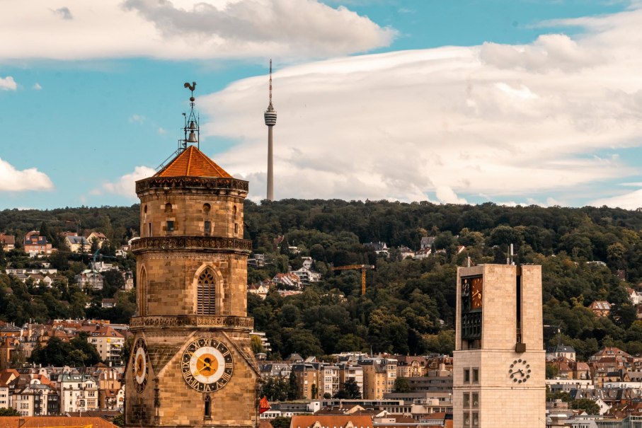 Die Stiftskirche in Stuttgart mit dem markanten Turm und der Uhr im Vordergrund. Rechts im Bild der Rathausturm. Im Hintergrund ist der Fernsehturm zu sehen, umgeben von gr&uuml;nen H&uuml;geln., &copy; Stuttgart-Marketing GmbH, Sarah Schmid