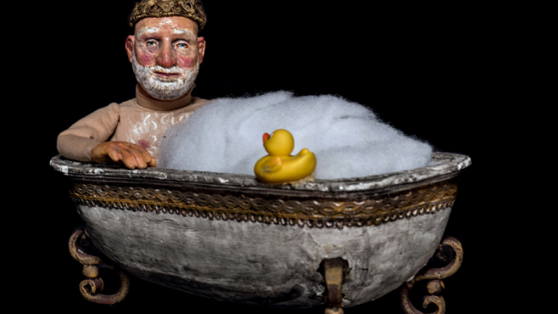 Eine Figur mit Krone sitzt in einer antiken Badewanne voller Schaum, neben einer gelben Gummiente., © Schäfer & Thieme Produktion Eine Figur mit Krone sitzt in einer antiken Badewanne voller Schaum, neben einer gelben Gummiente., © Schäfer & Thieme Produktion