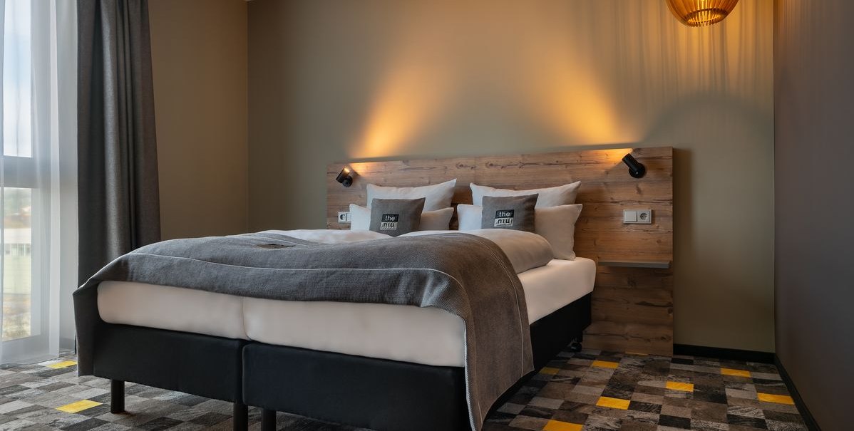 Modernes Hotelzimmer mit Doppelbett, Holzkopfteil, grauer Bettwäsche und stilvoller Beleuchtung. Der Teppich hat ein geometrisches Muster., © Novum Hospitality