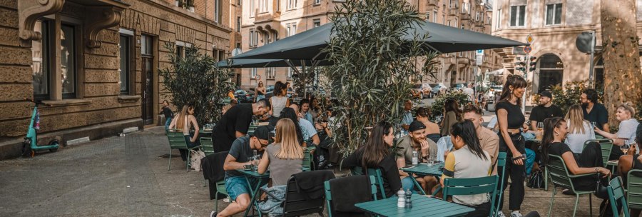 Menschen genie&szlig;en das Beisammensein an gr&uuml;nen Tischen im Freien vor einem Caf&eacute; in einer st&auml;dtischen Umgebung mit historischen Geb&auml;uden., &copy; Lupin, Stuttgart