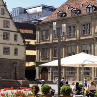 Der Schillerplatz in Stuttgart mit einer Statue im Vordergrund, umgeben von historischen Geb&auml;uden und Menschen, die unter Sonnenschirmen sitzen., &copy; Stuttgart-Marketing GmbH