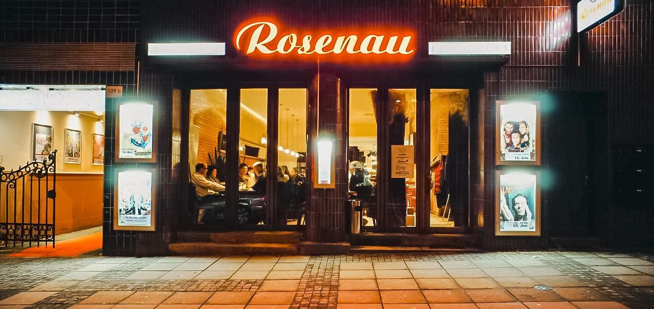 Eingang eines Lokals namens Rosenau mit leuchtendem Schild. Menschen sitzen drinnen, Plakate an den Wänden, gemütliche Atmosphäre., © Rosenau e. V., Stuttgart