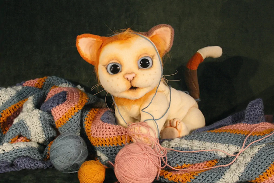 Eine Stoffkatze mit großen Augen sitzt auf einer bunten, gehäkelten Decke und spielt mit Wollknäueln in verschiedenen Farben., © FITHE – Das Figurentheater aus Ostbelgien Eine Stoffkatze mit großen Augen sitzt auf einer bunten, gehäkelten Decke und spielt mit Wollknäueln in verschiedenen Farben., © FITHE – Das Figurentheater aus Ostbelgien