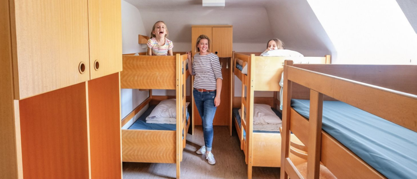 A quadruple room with two wooden bunk beds. Three people are smiling, two are standing on the beds, one is standing between them., &copy; DJH Baden-W&uuml;rttemberg e.V.