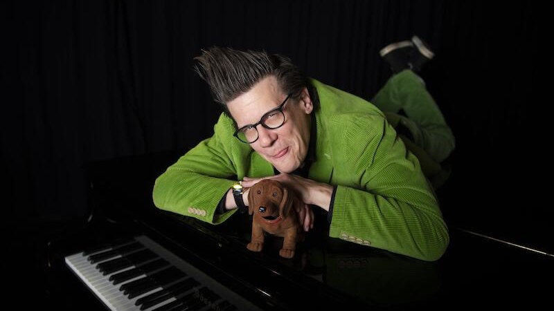 Person in gr&uuml;nem Anzug liegt l&auml;chelnd auf einem schwarzen Klavier, h&auml;lt ein Hundespielzeug. Dunkler Hintergrund., &copy; Renitenztheater Stuttgart e.V.