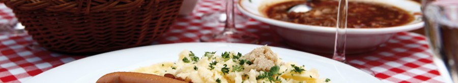 A plate of lentils, spaetzle and sausages on a red and white checkered tablecloth. A basket of bread and a glass of wine in the background., © Stuttgart-Marketing GmbH