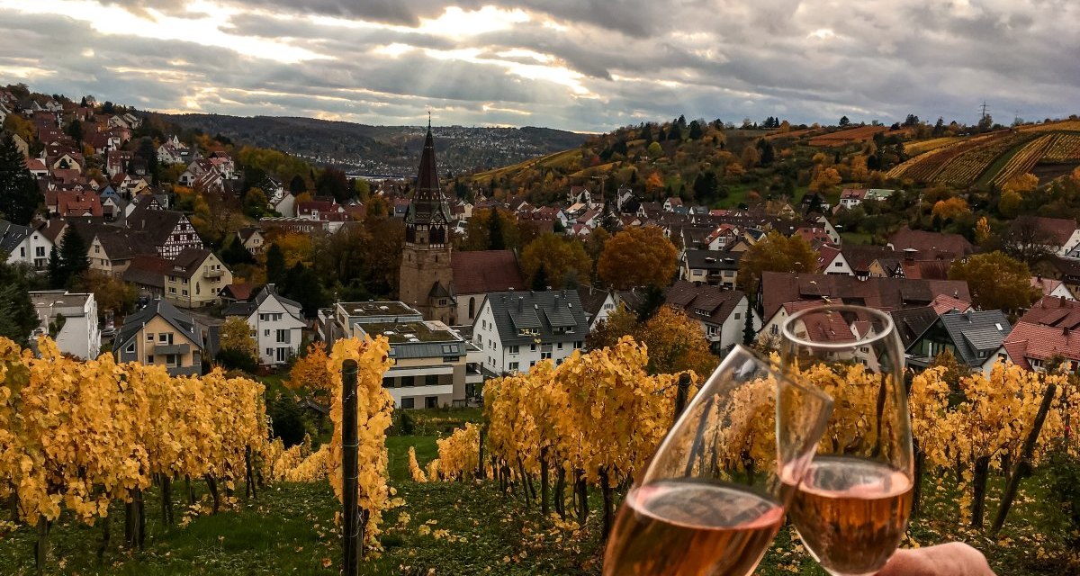 Zwei Personen stoßen mit Weingläsern in einem herbstlichen Weinberg an. Im Hintergrund ist eine Stadt mit Kirche und bunten Bäumen zu sehen., © Stuttgart-Marketing GmbH Zwei Personen stoßen mit Weingläsern in einem herbstlichen Weinberg an. Im Hintergrund ist eine Stadt mit Kirche und bunten Bäumen zu sehen., © Stuttgart-Marketing GmbH