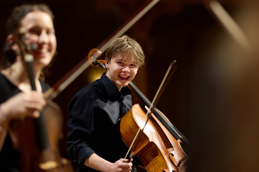 Ein junger Cellist lächelt während eines Auftritts auf der Bühne. Im Hintergrund ist eine weitere Musikerin mit einer Geige zu sehen., © Oliver Borchert Ein junger Cellist lächelt während eines Auftritts auf der Bühne. Im Hintergrund ist eine weitere Musikerin mit einer Geige zu sehen., © Oliver Borchert