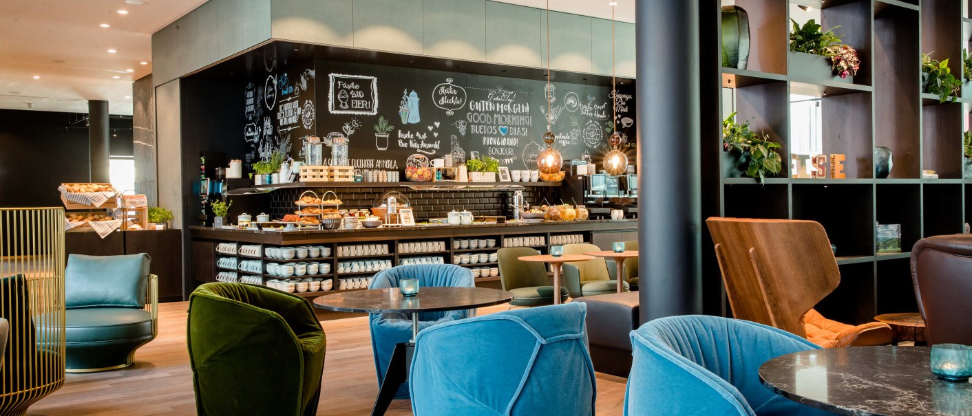 Modern lobby with colorful armchairs, tables and a coffee bar. The wall is decorated with chalk drawings. Plants and shelves complete the ambience., © Motel One - Stuttgart Hauptbahnhof Modern lobby with colorful armchairs, tables and a coffee bar. The wall is decorated with chalk drawings. Plants and shelves complete the ambience., © Motel One - Stuttgart Hauptbahnhof
