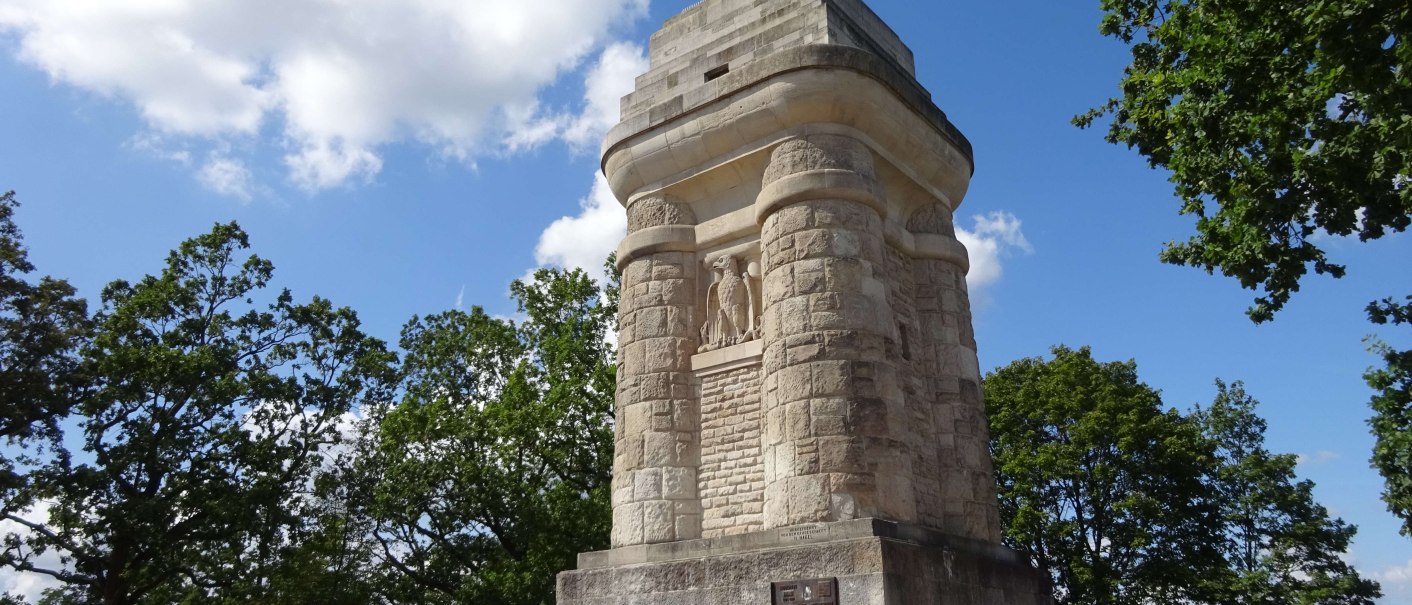 Ein massiver Bismarckturm aus Stein steht auf einem Platz, umgeben von Bäumen und unter einem blauen Himmel mit wenigen Wolken., © SMG Ein massiver Bismarckturm aus Stein steht auf einem Platz, umgeben von Bäumen und unter einem blauen Himmel mit wenigen Wolken., © SMG