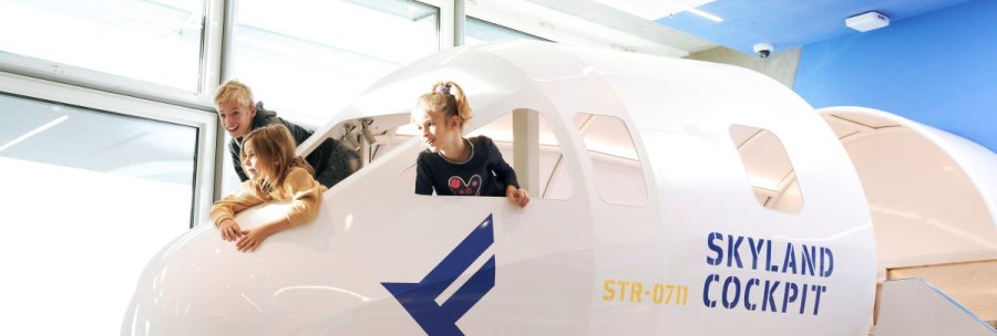 Drei Kinder spielen in einem Cockpit-Nachbau mit der Aufschrift 'SkyLand Cockpit' und blicken aus den Fenstern. Helle, moderne Umgebung., © Flughafen Stuttgart