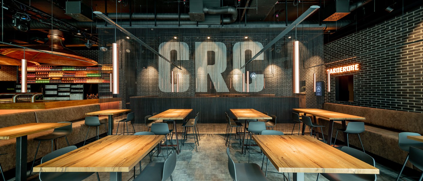 Modernes Café mit Holztischen, Stühlen und hängenden Lampen. "CRC" an der Wand, Flaschenregale im Hintergrund, Garderobe-Schild leuchtet., © Maks Richter