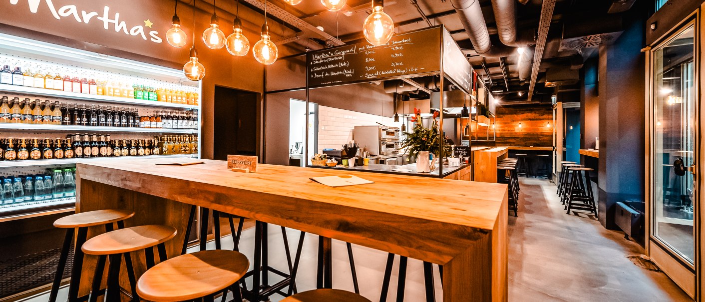 Modernes Café mit Holztischen, Hockern und hängenden Glühbirnen. Getränkekühlschrank und Menütafel im Hintergrund. Gemütliche Atmosphäre., © Andreas Dalferth Modernes Café mit Holztischen, Hockern und hängenden Glühbirnen. Getränkekühlschrank und Menütafel im Hintergrund. Gemütliche Atmosphäre., © Andreas Dalferth