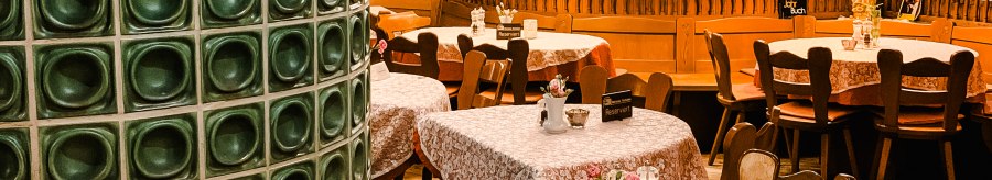 Cozy restaurant with wooden furniture, tablecloths and a green tiled stove. Lamps hanging from the ceiling, bar in the background., © Weinstube Kachelofen
