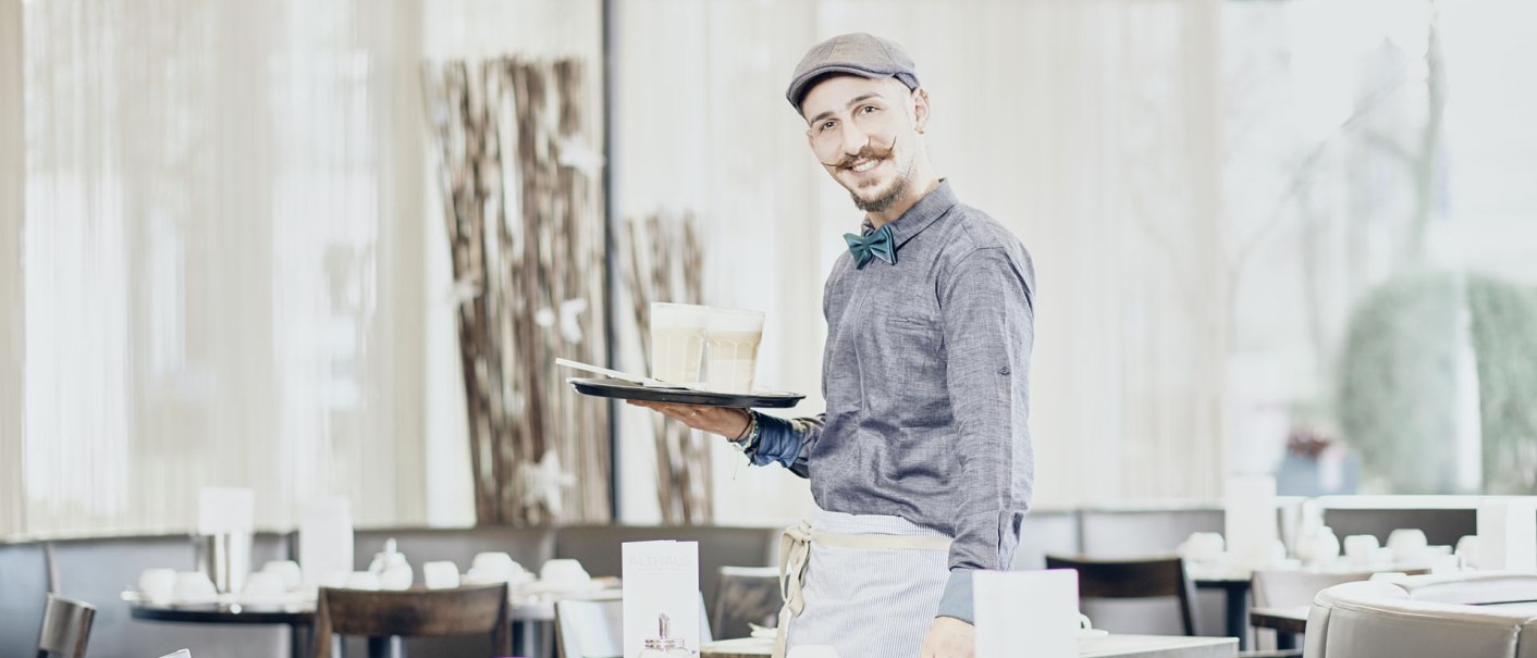 Ein lächelnder Kellner mit Mütze und Fliege serviert Getränke in einem hellen, modernen Frühstücksbereich., © MASEVEN Stuttgart SI-Centrum