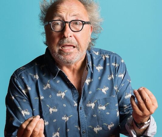 A man with disheveled hair and glasses gesticulates animatedly against a blue background., &copy; Theaterhaus Stuttgart e.V.