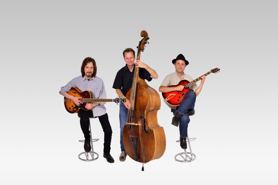 Ein Jazztrio mit zwei Gitarristen und einem Kontrabassisten posiert vor einem grauen Hintergrund. Alle sitzen auf Hockern., &copy; Saite16