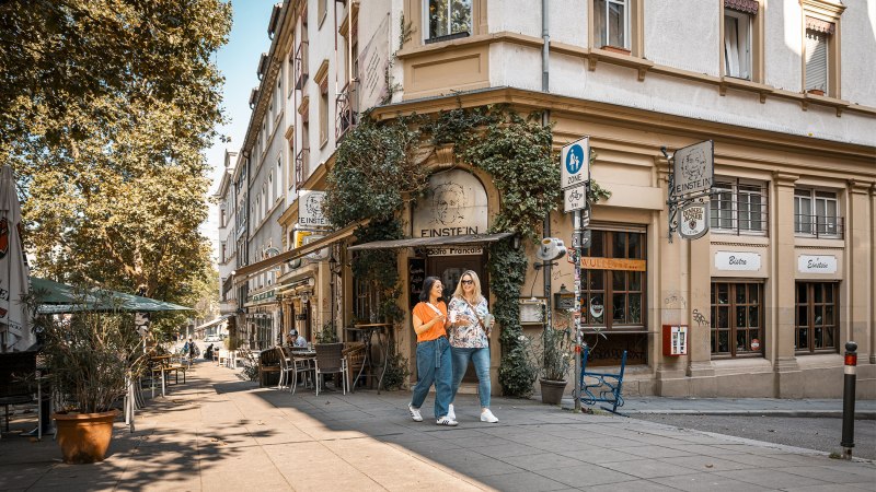 Zwei Frauen spazieren lächelnd am Café Einstein vorbei. Die Straße ist gesäumt von Bäumen und Außensitzplätzen. Es herrscht eine entspannte Atmosphäre., © Stuttgart-Marketing GmbH, Sarah Schmid