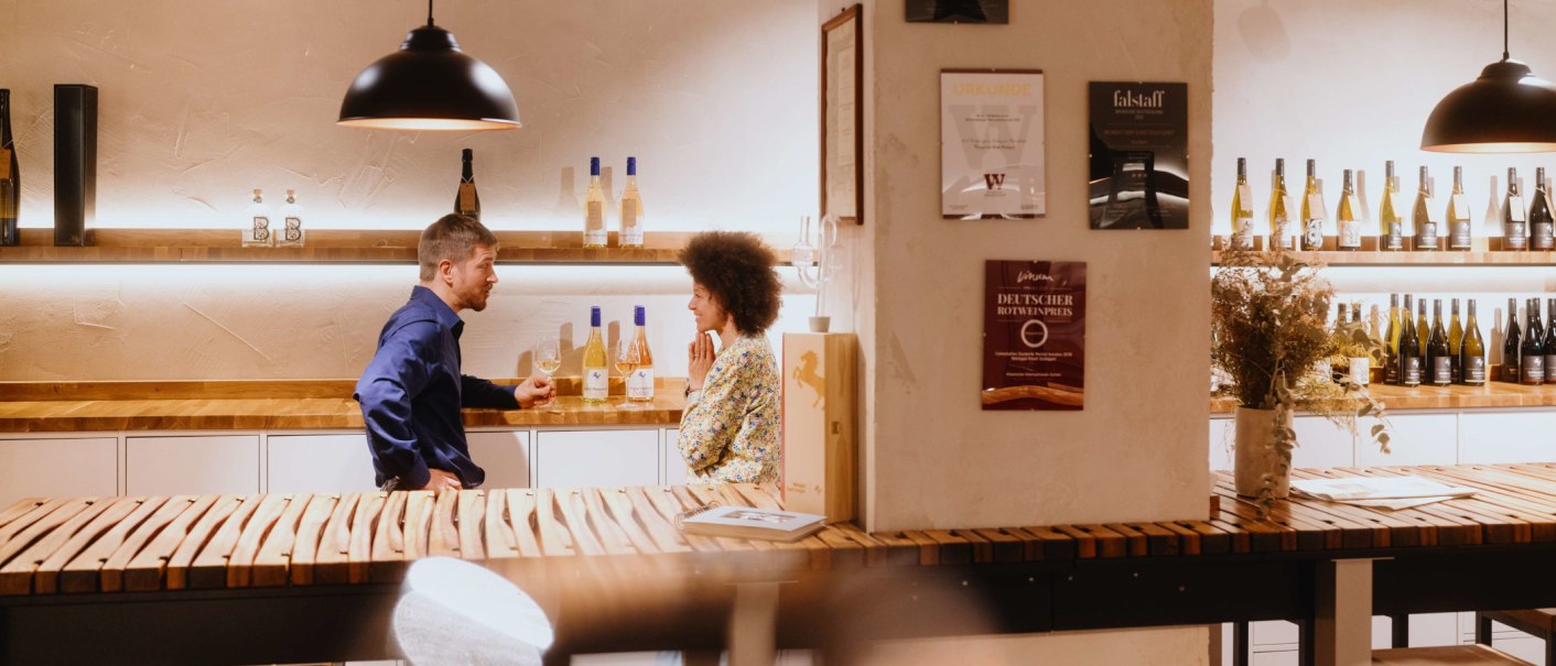 Zwei Personen unterhalten sich in einer Vinothek. Weinflaschen stehen auf Regalen, Urkunden an der Wand. Gemütliche Atmosphäre mit Holztischen., © SMG, Alwin Maigler Zwei Personen unterhalten sich in einer Vinothek. Weinflaschen stehen auf Regalen, Urkunden an der Wand. Gemütliche Atmosphäre mit Holztischen., © SMG, Alwin Maigler