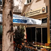 Außenbereich eines Cafés namens Oblomow mit Holztischen, Stühlen und einem Gauloises-Sonnenschirm. Ein Baum steht im Vordergrund., © SMG_Sarah Schmid Außenbereich eines Cafés namens Oblomow mit Holztischen, Stühlen und einem Gauloises-Sonnenschirm. Ein Baum steht im Vordergrund., © SMG_Sarah Schmid