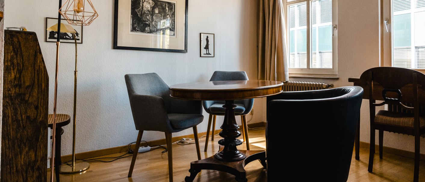 Ein gemütlicher Raum mit Holzbalken, einem runden Tisch, verschiedenen Stühlen und Kunstwerken an der Wand. Sonnenlicht fällt durch die Fenster., © SMG Stuttgart Marketing GmbH - Sarah Schmid