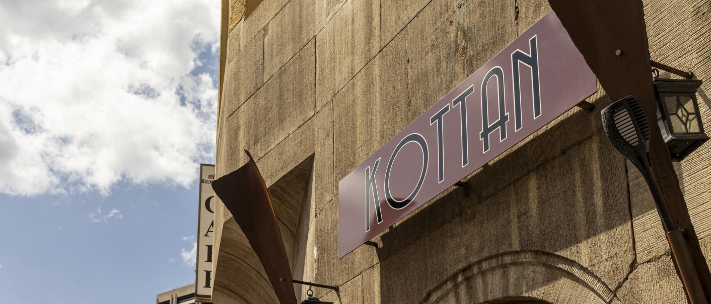 Ein Schild mit der Aufschrift 'KOTTAN' hängt an einer Gebäudewand unter einem blauen Himmel mit Wolken., © SMG, Sarah Schmid