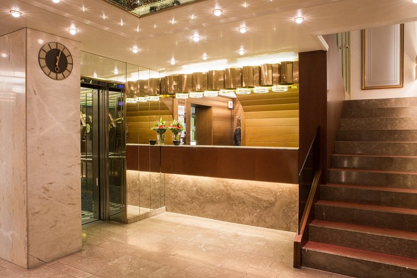 Elegante Hotelrezeption mit Marmorboden, Uhr an der Wand, Aufzug und Treppe. Der Empfangstresen ist mit Blumen dekoriert., © Hotel Royal