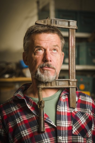 Ein Mann mit grauem Bart und kariertem Hemd hält eine große Schraubzwinge um seinen Kopf. Er blickt nach oben, im Hintergrund sind Werkzeuge zu sehen., © Rosenau Kultur e.V. Ein Mann mit grauem Bart und kariertem Hemd hält eine große Schraubzwinge um seinen Kopf. Er blickt nach oben, im Hintergrund sind Werkzeuge zu sehen., © Rosenau Kultur e.V.