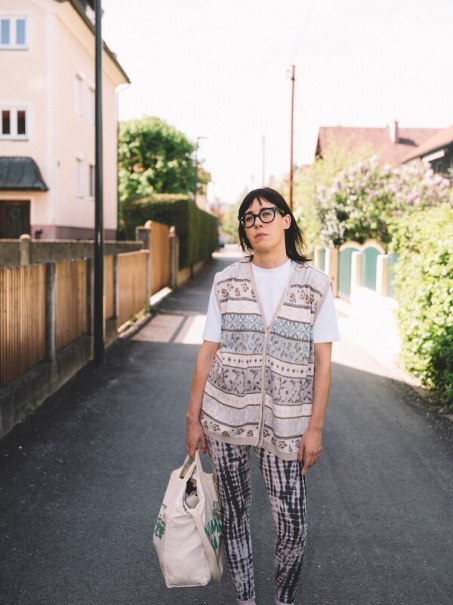 Person mit Brille und gemusterter Weste steht auf einer ruhigen Straße mit Einkaufstasche. Im Hintergrund sind Häuser und Büsche zu sehen., © Rosenau Kultur e.V.
