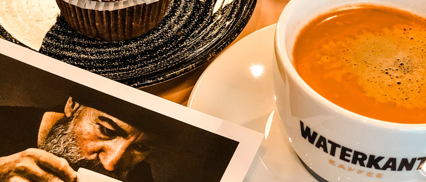 Cupcake mit Schokocreme und Nüssen, Kaffee in Tasse mit 'Waterkant'-Logo, Zuckerstreuer und Foto eines Mannes mit Kaffeetasse auf Tisch., © Sanija Göhner