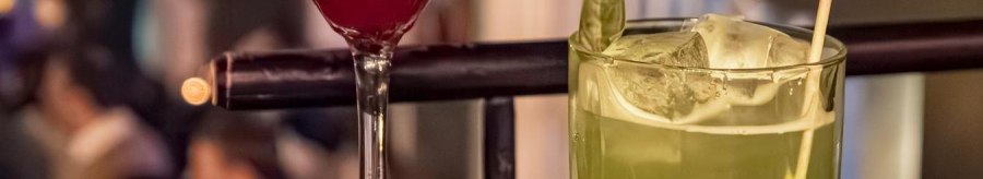 Two cocktails on a bar: a red one in a martini glass and a green one in a tumbler glass with a straw, blurred bar scene in the background., © SMG Stuttgart Marketing GmbH - Sarah Schmid