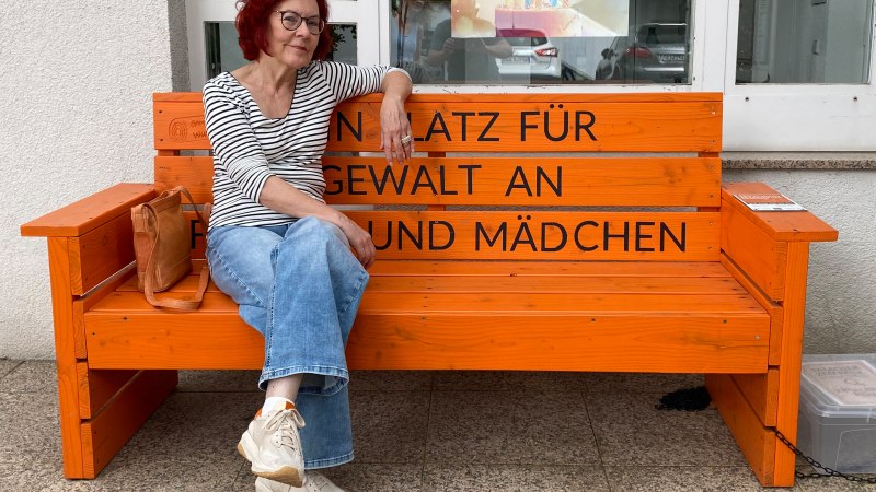 Eine Frau mit rotem Haar und Brille sitzt auf einer orangefarbenen Bank mit der Aufschrift 'Kein Platz f&uuml;r Gewalt an Frauen und M&auml;dchen'., &copy; Foto: T. Faupel